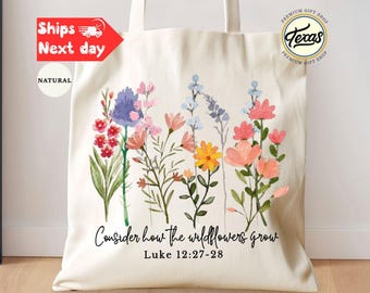 May include: A natural canvas tote bag featuring a watercolor floral design in shades of pink, purple, yellow, and blue. The text "Consider how the wildflowers grow" and Bible verse Luke 12:27-28 are printed below the flowers. The bag has two handles.
