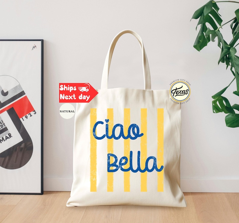 Op de afbeelding: Cr&egrave;mekleurige draagtas met verticale gele strepen en de woorden "Ciao Bella" in blauw schrift. De tas heeft lange handvatten en een "Texas Premium Gift Shop"-logo. Een ingelijste kunstdruk en groen zijn op de achtergrond.