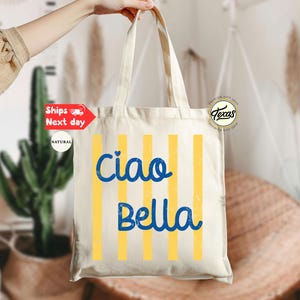 Op de afbeelding: Cr&egrave;mekleurige draagtas met verticale gele strepen en de woorden "Ciao Bella" in blauw schrift. De tas wordt door een hand omhoog gehouden. De achtergrond omvat planten en een rotan stoel.