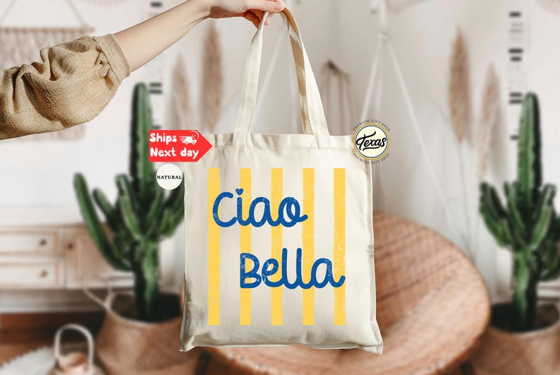 Op de afbeelding: Cr&egrave;mekleurige draagtas met verticale gele strepen en de woorden "Ciao Bella" in blauw schrift. De tas wordt door een hand omhoog gehouden. De achtergrond omvat planten en een rotan stoel.