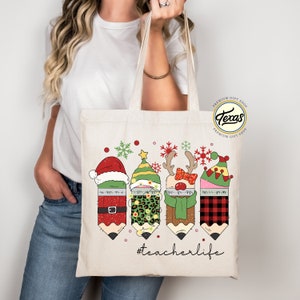 May include: A white canvas tote bag with a Christmas-themed design featuring four pencils dressed as Santa, an elf, a reindeer, and a snowman. The pencils have glitter and are wearing festive hats and accessories. The text "#teacherlife" is printed below the pencils.