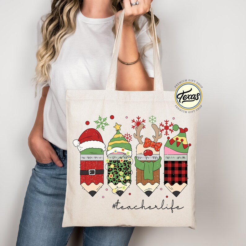 May include: A white canvas tote bag with a Christmas-themed design featuring four pencils dressed as Santa, an elf, a reindeer, and a snowman. The pencils have glitter and are wearing festive hats and accessories. The text "#teacherlife" is printed below the pencils.