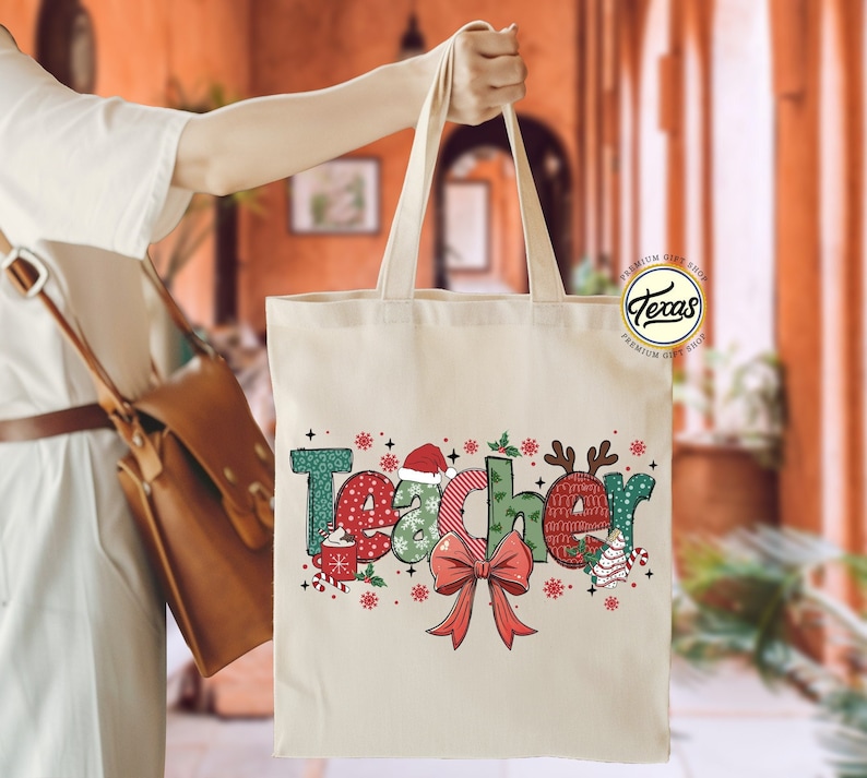 Puede incluir: Una bolsa de lona blanca con un dise&ntilde;o navide&ntilde;o que dice "Teacher" en letras de colores con decoraciones festivas. La bolsa tiene un lazo rojo y un reno verde y rojo.