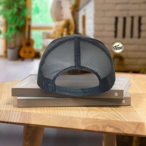 May include: A black and gray mesh trucker hat with a black adjustable strap. The hat is sitting on top of two books.