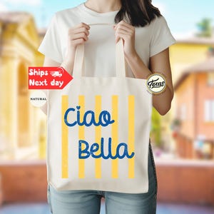 Op de afbeelding: Een natuurlijke canvas draagtas met verticale gele strepen en de woorden "Ciao Bella" in blauw schrift. De tas wordt door een persoon vastgehouden. De achtergrond is wazig.