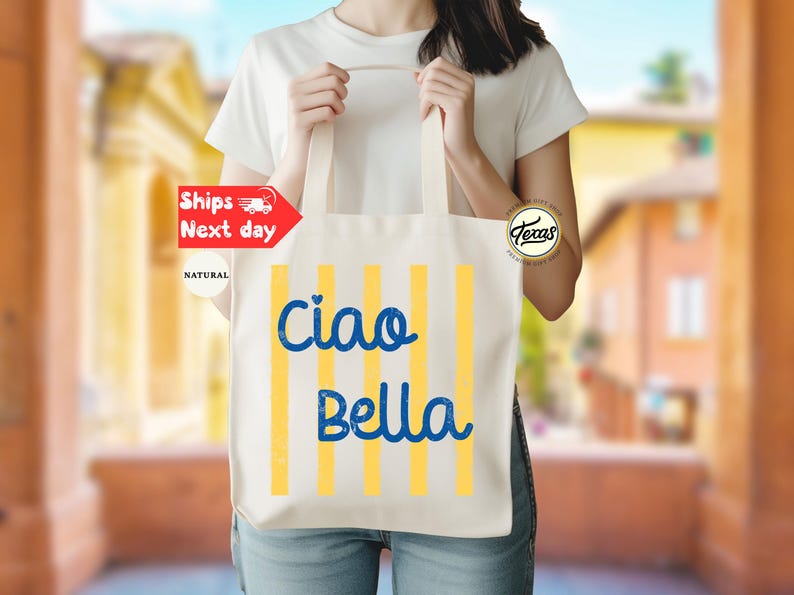 Op de afbeelding: Een natuurlijke canvas draagtas met verticale gele strepen en de woorden "Ciao Bella" in blauw schrift. De tas wordt door een persoon vastgehouden. De achtergrond is wazig.