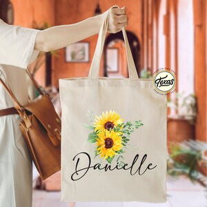 K&ouml;nnte beinhalten: Eine wei&szlig;e Canvas-Tragetasche mit einem Sonnenblumen-Design und dem Namen "Danielle" in schwarzer Schrift. Die Tasche hat einen braunen Ledergriff und ein kleines rundes Logo mit dem Text "Texas Premium Gift Shop".