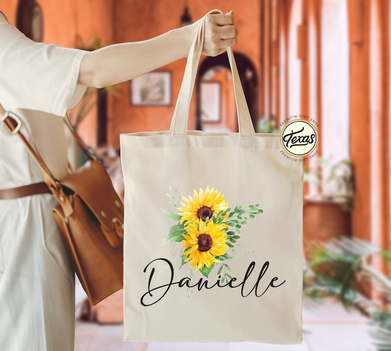 K&ouml;nnte beinhalten: Eine wei&szlig;e Canvas-Tragetasche mit einem Sonnenblumen-Design und dem Namen "Danielle" in schwarzer Schrift. Die Tasche hat einen braunen Ledergriff und ein kleines rundes Logo mit dem Text "Texas Premium Gift Shop".