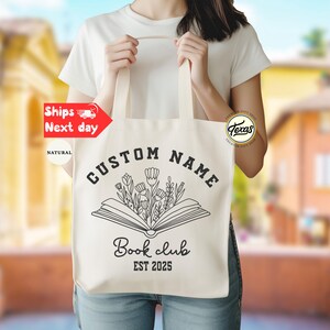 May include: Natural canvas tote bag with black text and a floral book design. The text reads "CUSTOM NAME" above an open book with flowers, and "Book club EST 2025" below. The bag has long handles.