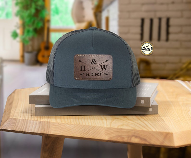 May include: A dark blue trucker hat with a brown leather patch that says "H & W 01.12.2023" with two arrows crossing in the middle.