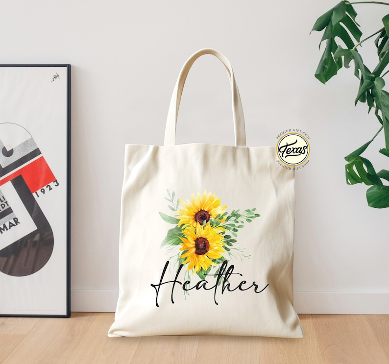 K&ouml;nnte beinhalten: Eine wei&szlig;e Canvas-Tragetasche mit einem Blumendesign aus Sonnenblumen und Gr&uuml;n. Die Tasche tr&auml;gt den Namen "Heather" in schwarzer, kursiv geschriebener Schrift.