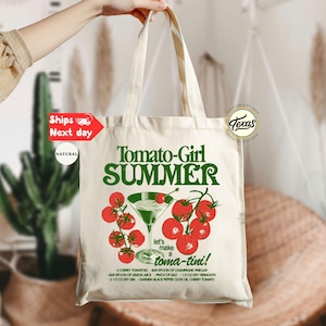 Peut inclure: Un sac fourre-tout en toile naturelle avec l'inscription "Tomato-Girl SUMMER" en vert, orn&eacute; d'une illustration de cocktail &agrave; la tomate. Le sac comprend &eacute;galement une recette de "toma-tini".