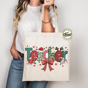 Puede incluir: Una bolsa de lona blanca con un dise&ntilde;o navide&ntilde;o que dice "Teacher" en letras de colores con decoraciones festivas. La bolsa tiene un lazo rojo y un peque&ntilde;o logotipo circular que dice "Premium Gift Shop Texas".