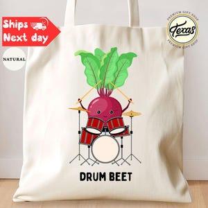 May include: A natural canvas tote bag featuring a cartoon beet playing a drum set. The beet has a smiling face, green leafy hair, and is holding drumsticks. The text "DRUM BEET" is printed below the drum set.