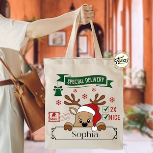 May include: A beige canvas tote bag with a Christmas-themed design. The design includes a reindeer wearing a Santa hat, snowflakes, and the text "Special Delivery" and "2X NICE". The name "Sophia" is printed below the reindeer.