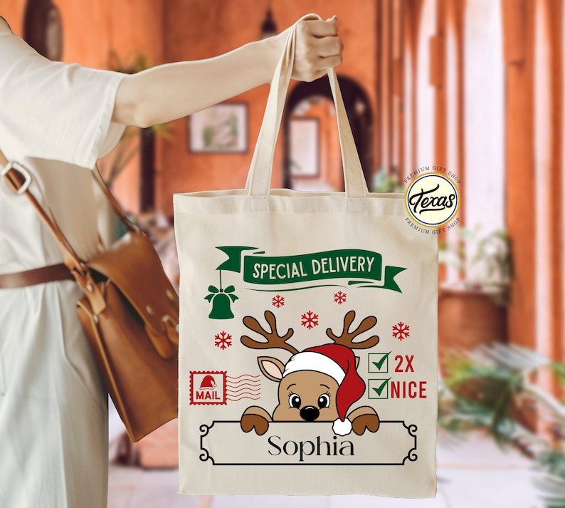 May include: A beige canvas tote bag with a Christmas-themed design. The design includes a reindeer wearing a Santa hat, snowflakes, and the text "Special Delivery" and "2X NICE". The name "Sophia" is printed below the reindeer.