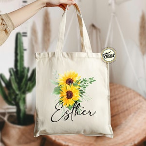 K&ouml;nnte beinhalten: Eine wei&szlig;e Canvas-Tragetasche mit einem Blumendesign aus Sonnenblumen und Gr&uuml;n. Die Tasche hat einen schwarzen Griff und den Namen "Esther" in schwarzer Kursivschrift.