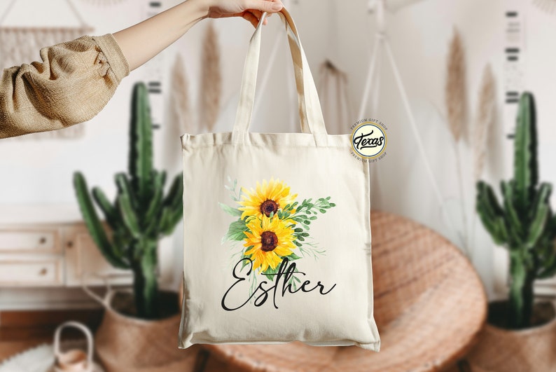K&ouml;nnte beinhalten: Eine wei&szlig;e Canvas-Tragetasche mit einem Blumendesign aus Sonnenblumen und Gr&uuml;n. Die Tasche hat einen schwarzen Griff und den Namen "Esther" in schwarzer Kursivschrift.