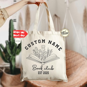 May include: Cream-colored tote bag with black text and a floral book design. The text reads "CUSTOM NAME" above an open book with flowers, and "Book club EST 2025" below. The bag is held up by a person.