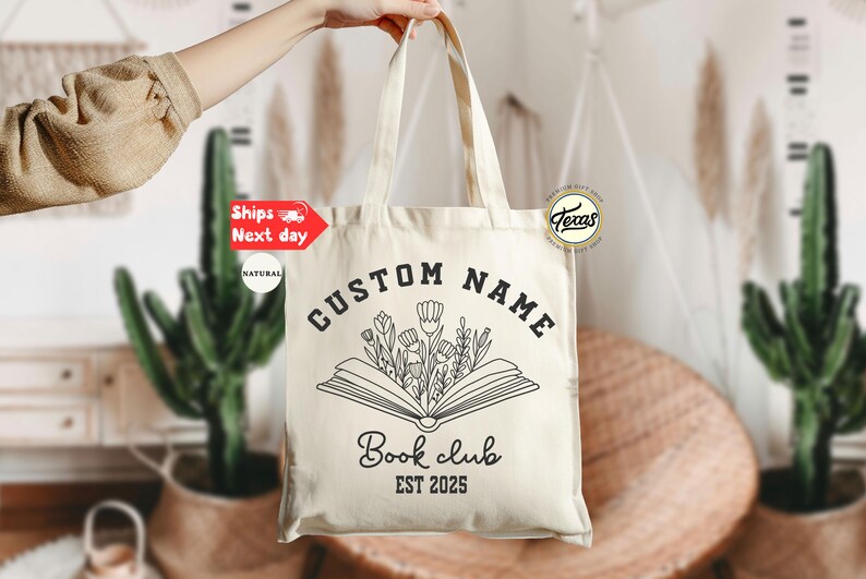 May include: Cream-colored tote bag with black text and a floral book design. The text reads "CUSTOM NAME" above an open book with flowers, and "Book club EST 2025" below. The bag is held up by a person.