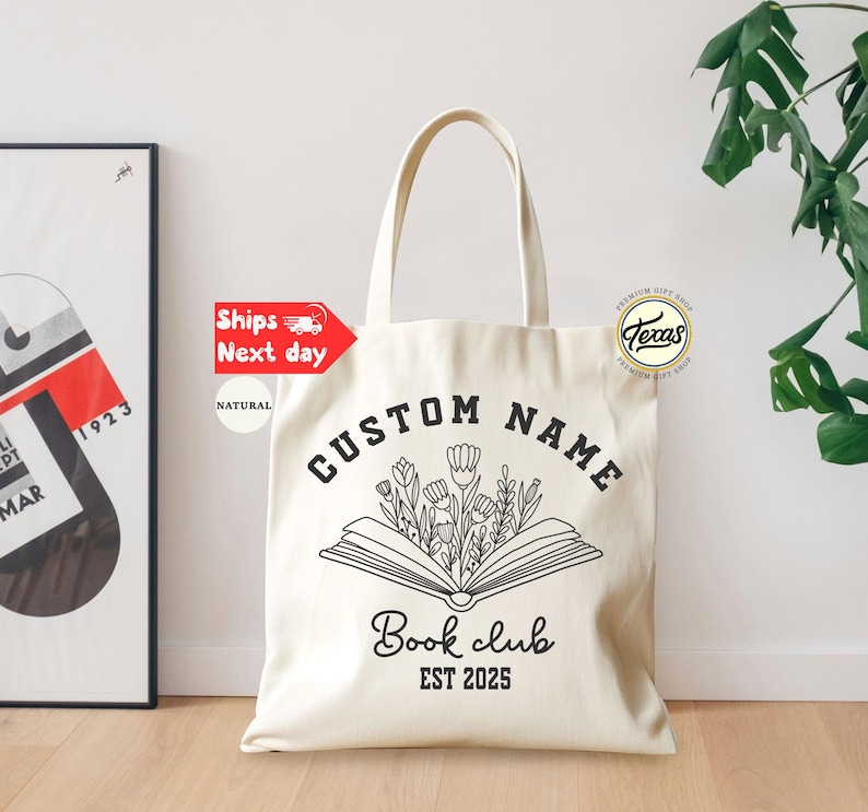May include: Cream-colored tote bag with black text and a floral book design. The bag reads "CUSTOM NAME", "Book club", and "EST 2025". A framed art print and a plant are in the background.