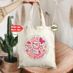 May include: A white canvas tote bag with a floral design and the word "madre" in the center. The design is surrounded by a circle of words in Spanish, including "comprendente", "protectora", "confiada", and "amable".