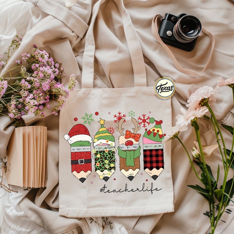 May include: A white canvas tote bag with a Christmas-themed design featuring four pencils dressed as Santa, an elf, a reindeer, and a snowman. The pencils are surrounded by snowflakes and red dots. The text "#teacherlife" is printed below the pencils.