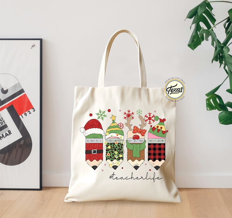 May include: A white canvas tote bag with a Christmas-themed design featuring four pencils dressed as Santa, an elf, a reindeer, and a snowman. The pencils are wearing festive hats and scarves. The text "#teacherlife" is printed below the pencils.