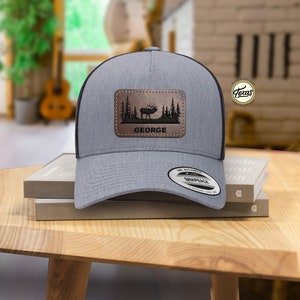 May include: A gray and black trucker hat with a brown leather patch featuring a silhouette of an elk in a forest scene. The patch also includes the name "GEORGE".
