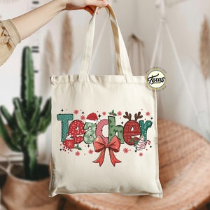 Puede incluir: Una bolsa de lona blanca con un dise&ntilde;o navide&ntilde;o que dice "Teacher" en letras de colores con un lazo rojo y copos de nieve.