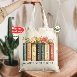 May include: Cream-colored tote bag featuring a floral design above a row of books. Each book spine has a name and a cross symbol. The text "WOMEN OF THE BIBLE" is printed below the books. The bag is held by a person.
