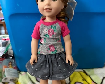 Op de afbeelding: Een pop met rossig haar, groene ogen en sproeten, gekleed in een grijs en roze shirt met bloemenprint, een spijkerbroekrok met witte stippen en een zilveren strik. De tekst op de blauwe container luidt "A Girl & Nellie Wishes".