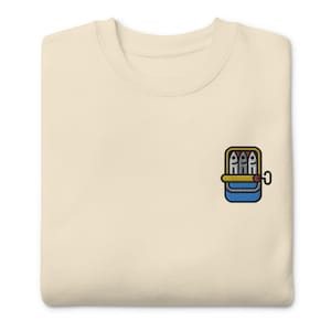 May include: A cream-colored t-shirt with an embroidered image of a can of sardines. The can is yellow with a blue lid and a red pull tab. Three sardines are visible inside the can.