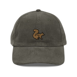 Peut inclure: Casquette de baseball en velours côtelé vert olive avec un emblème d'écureuil marron. La casquette a une visière incurvée et une sangle réglable. Le motif de l'écureuil est brodé sur le devant. Un accessoire décontracté.