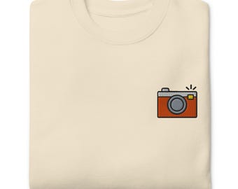 May include: A cream colored t-shirt with a red and grey camera embroidered on the front. The camera has a yellow flash.