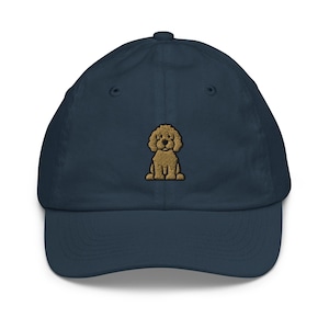 May include: A navy blue baseball cap featuring an embroidered image of a light brown dog. The cap has a curved brim and an adjustable strap at the back. The dog is sitting and has a fluffy appearance.