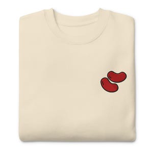 May include: A cream-coloured sweatshirt with a crew neck. The sweatshirt has an embroidered design of two red kidney beans with black outlines. The fabric appears soft and comfortable.