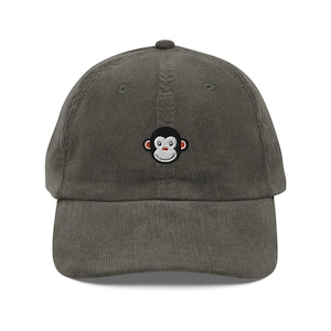 May include: A corduroy baseball cap in a muted olive green color. The cap features a small embroidered monkey face on the front. The monkey has a black outline, white face, and red and orange details. The cap has a curved brim and a rounded top.
