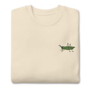 May include: A light beige t-shirt with a green embroidered grasshopper on the front.