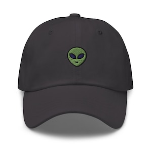 Puede incluir: Una gorra de béisbol gris oscuro con un rostro de extraterrestre verde bordado con ojos azules.
