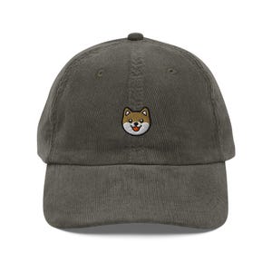 May include: A corduroy baseball cap in a muted olive green color. The cap features a small embroidered Shiba Inu dog face on the front, with brown, white, and black details. The dog has a happy expression with its tongue sticking out.