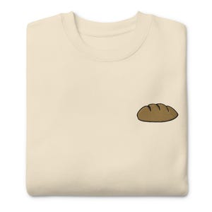 May include: Cream-colored sweatshirt with a small embroidered loaf of brown bread on the front. The bread has a dark brown outline and detailed stitching. The sweatshirt has a crew neck and a relaxed fit.