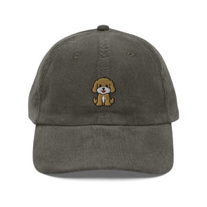 May include: An olive green corduroy baseball cap with an embroidered cartoon puppy. The puppy has tan fur, a white face, and a red tongue. The cap has a curved peak and a fabric-covered button on top.