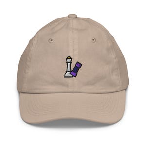 Puede incluir: Gorra de béisbol beige con visera curva. La gorra presenta un diseño bordado de una pieza de ajedrez blanca y una pieza de ajedrez morada. La gorra tiene un ajuste estructurado y un cierre de correa de tela.