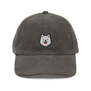 Può includere: Cappellino da baseball in velluto a coste verde oliva. Presenta una piccola toppa ricamata di un cane bianco sorridente con la lingua rossa. Il cappello ha una visiera curva e un cinturino regolabile.