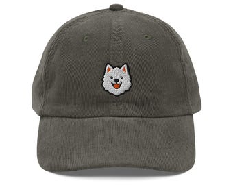 May include: A corduroy baseball cap in a muted green color. The cap features a small embroidered patch of a smiling white dog with a red tongue. The hat has a curved brim and an adjustable strap.