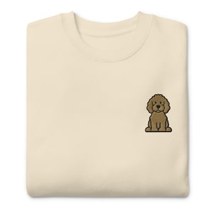 May include: Cream-colored sweatshirt with a small embroidered brown poodle design on the chest. The sweatshirt has a crew neck and a relaxed fit, suitable for casual wear. The poodle is depicted in a seated position.