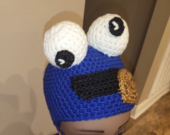 May include: Blue crocheted hat with white and black eyes and a brown cookie design. The hat is worn on a mannequin head.