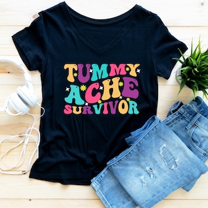 May include: A black t-shirt with a colorful graphic that says "Tummy Ache Survivor". The shirt is laid flat on a wooden surface with a pair of blue jeans and a white pair of headphones.