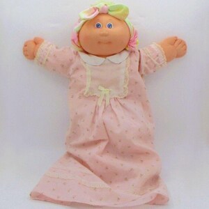 May include: A vintage doll with a peach-coloured face, blue eyes, and a floral head wrap. The doll wears a pink floral-print dress with lace trim and a white collar. The doll's arms are outstretched, and the overall appearance is soft and nostalgic.